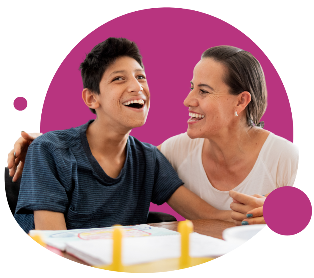 A boy wearing a dark grey top is sitting at a desk with a woman who is smiling at him. They both appear to be smiling and are happy. The image is placed on top of a pink circle.