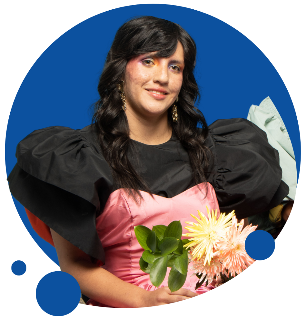 A person in a black and pink formal dress is wearing colourful makeup and smiling at the camera. The image is placed on top of a blue circle. They are holding a bouquet of white flowers.