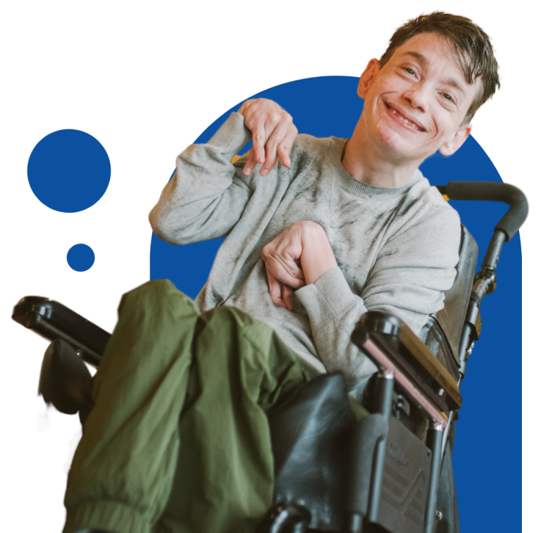 A person sitting in a wheelchair has short brown hair, a light gray jumper and khaki green pants on. They are smiling at the camera. The image is placed on top of a blue coloured arch-like shape.